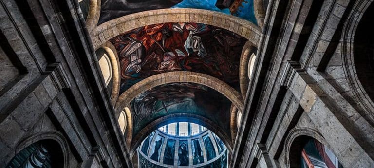 The image captures the stunning frescoes and arches inside the Hospicio Cabañas, showcasing vibrant colors and intricate artwork beneath its historical dome.