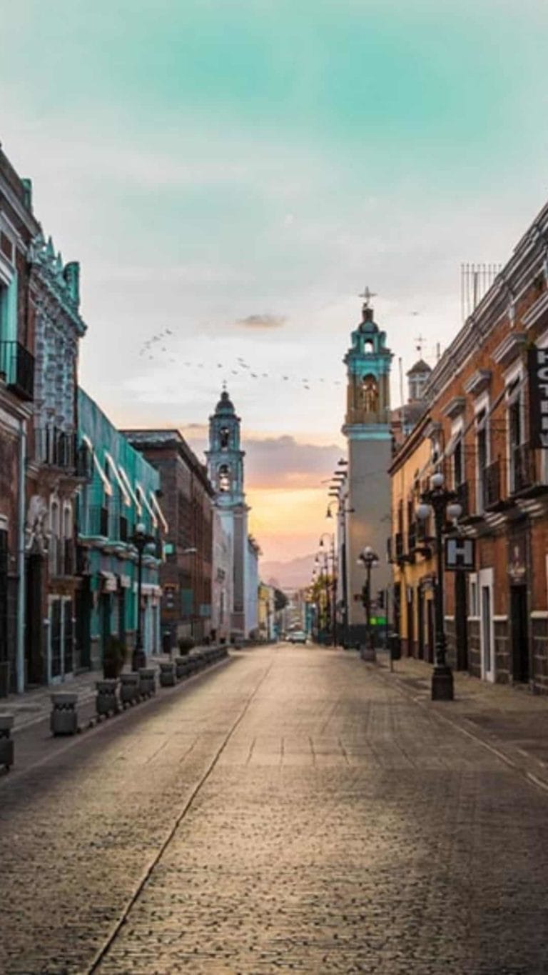 A serene, empty street with colonial architecture leads to Puebla Cathedral towers, under a vibrant sky with birds in flight at sunset.