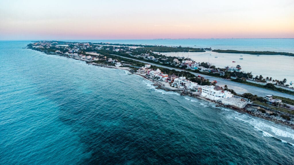 Coastal view of Isla Mujeres, Mexico, with colorful houses lining the shoreline and a tranquil sea under a soft pastel sunset sky.