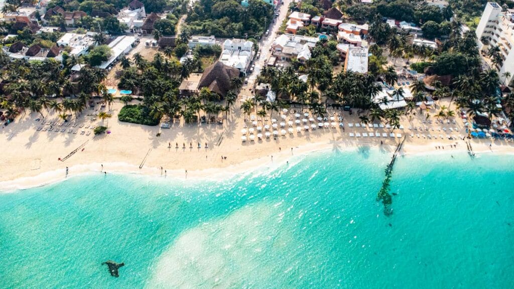 Aerial view of a vibrant tropical beach scene with turquoise waters, lined with palm trees, sun loungers, and people enjoying the shoreline.
