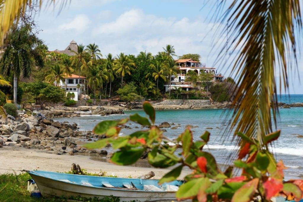 Peaceful beachfront framed by tropical trees and a rocky shoreline, with villas and palm trees dotting the coast, viewed through lush green leaves.