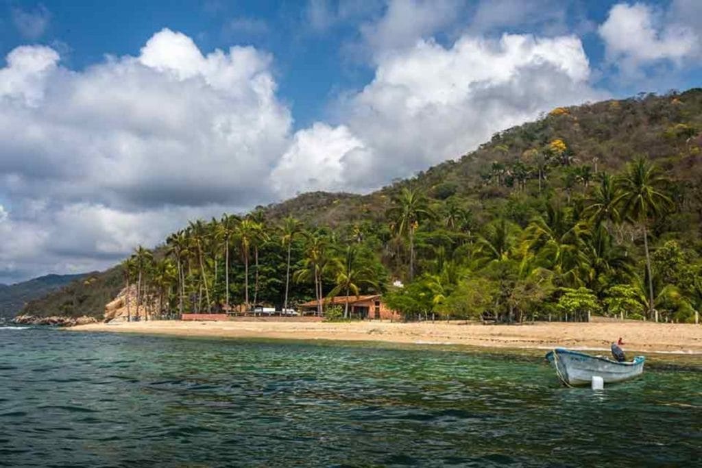 View of Quimixto Beach as seen from the water, with a rustic beachfront house surrounded by tall palm trees and backed by a lush hillside under partly cloudy skies.