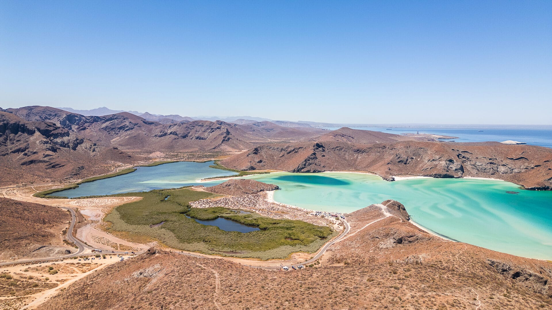 Aerial view of Balandra Beach, featuring turquoise waters, surrounding arid mountains, and sparse vegetation under clear, sunny skies. Cactus plants visible on land.