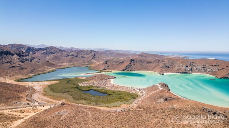Aerial view of Balandra Beach with turquoise waters, surrounded by rugged desert hills under a clear blue sky. Cars parked in the area.
