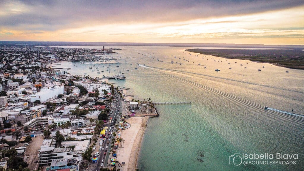Aerial view of La Paz coastline with numerous boats, a bustling cityscape, and scenic shoreline, under a colorful sunset sky.