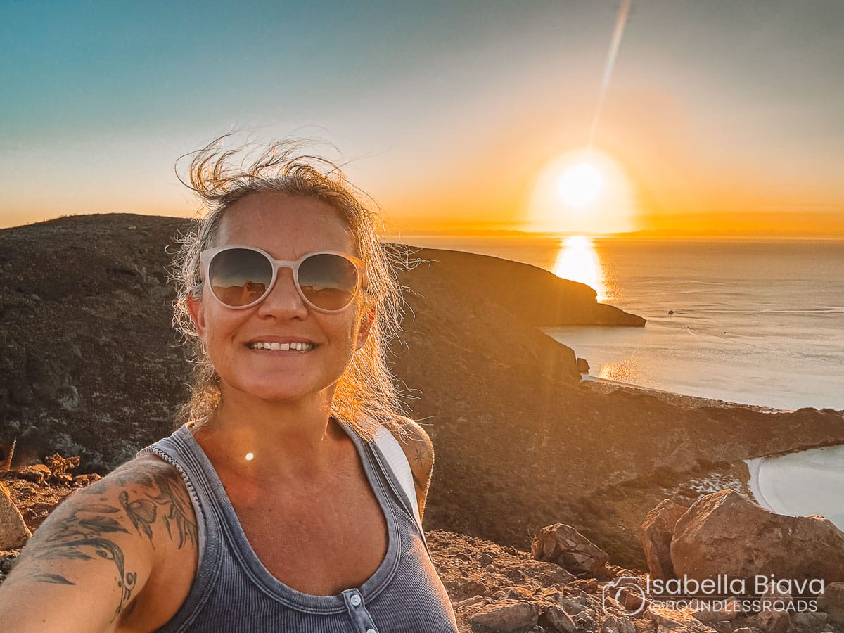 A person wearing sunglasses smiles during sunset on a rocky cliff overlooking a calm ocean and distant headland.