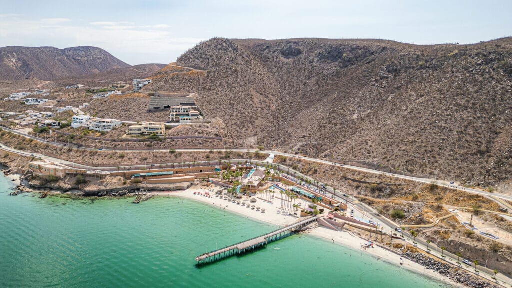 Coastal landscape with sandy beach, vibrant umbrellas, and a long pier. Hillside houses overlook the blue-green sea. Desert mountains in the background.