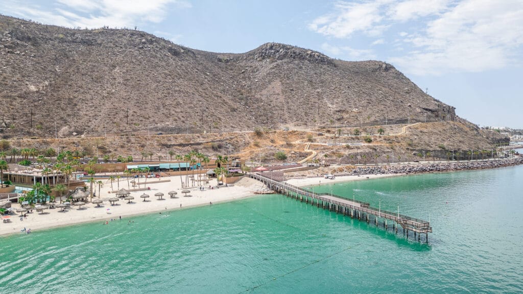 A tranquil beach scene with a wooden pier, palm trees, and people enjoying the clear turquoise water, surrounded by rocky hills.