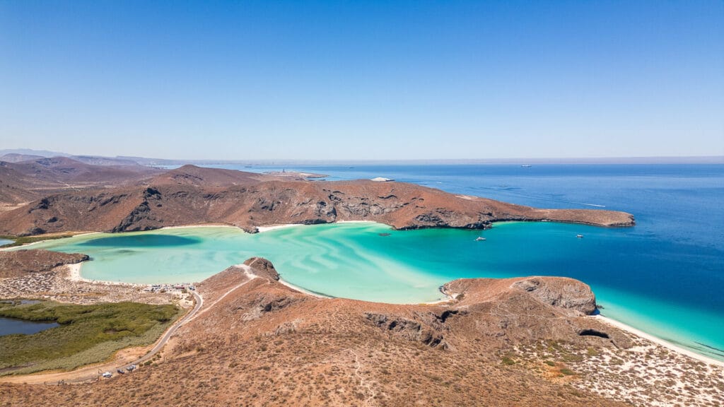 Aerial view of Balandra Beach, featuring turquoise waters and rocky hills under a clear blue sky. Boats are anchored in the bay.