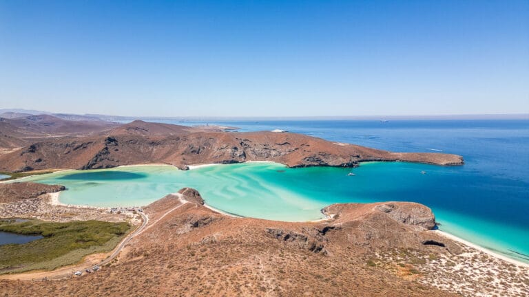 Aerial view of Balandra Beach, featuring turquoise waters and rocky hills under a clear blue sky. Boats are anchored in the bay.