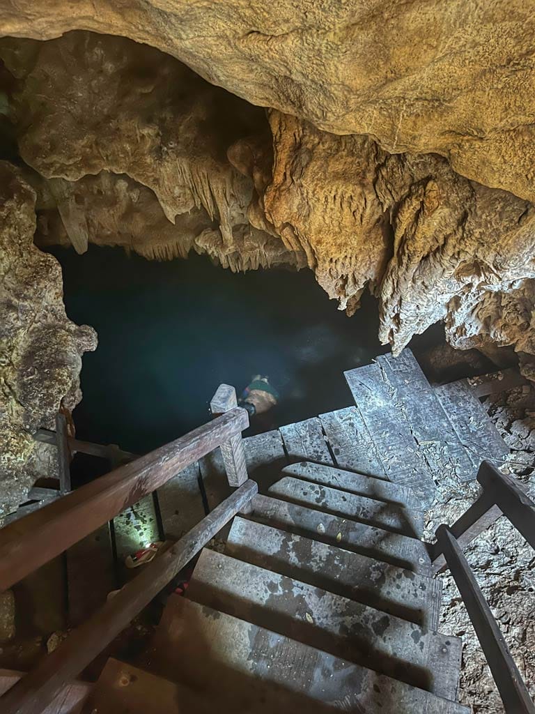 A person swims in a cave pool with rocky formations and wooden stairs descending into the water, creating a serene, natural setting.