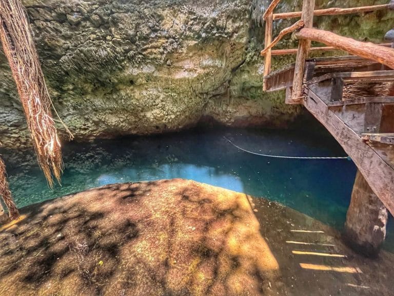 A serene, turquoise water cenote with overhanging vines and a rustic wooden platform surrounded by natural rock formations.