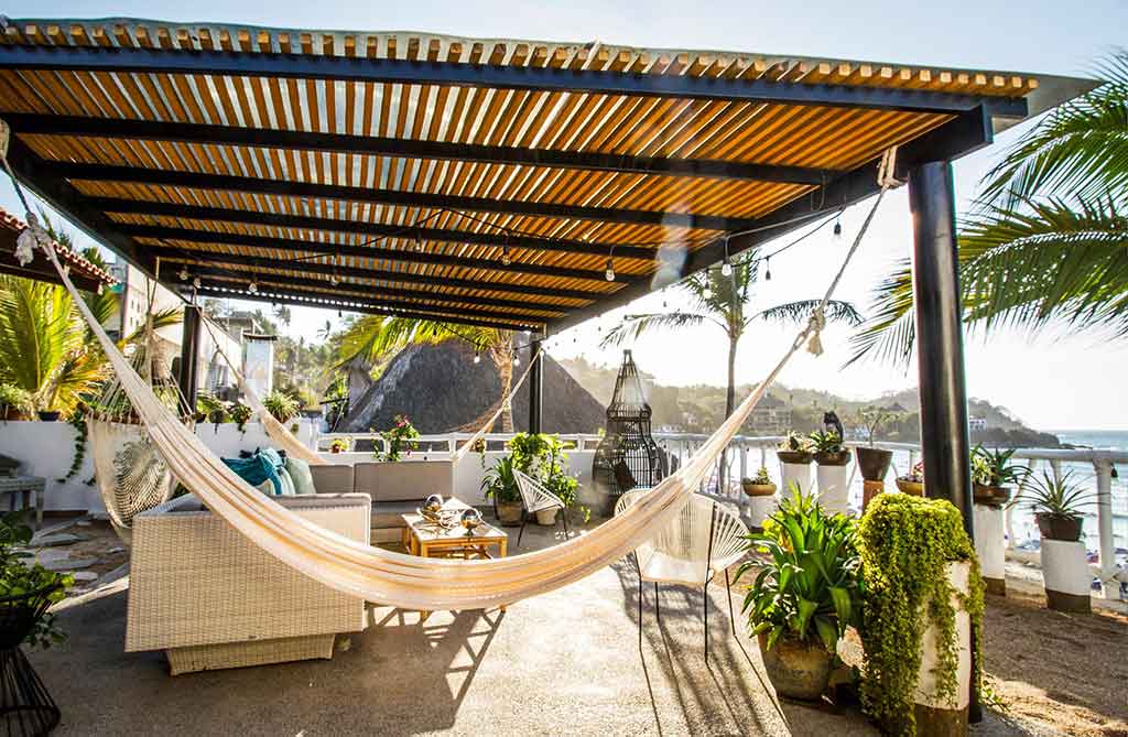 Sunlit rooftop lounge overlooking the ocean, featuring a hammock, tropical plants, cozy seating, and a wooden pergola for shade.