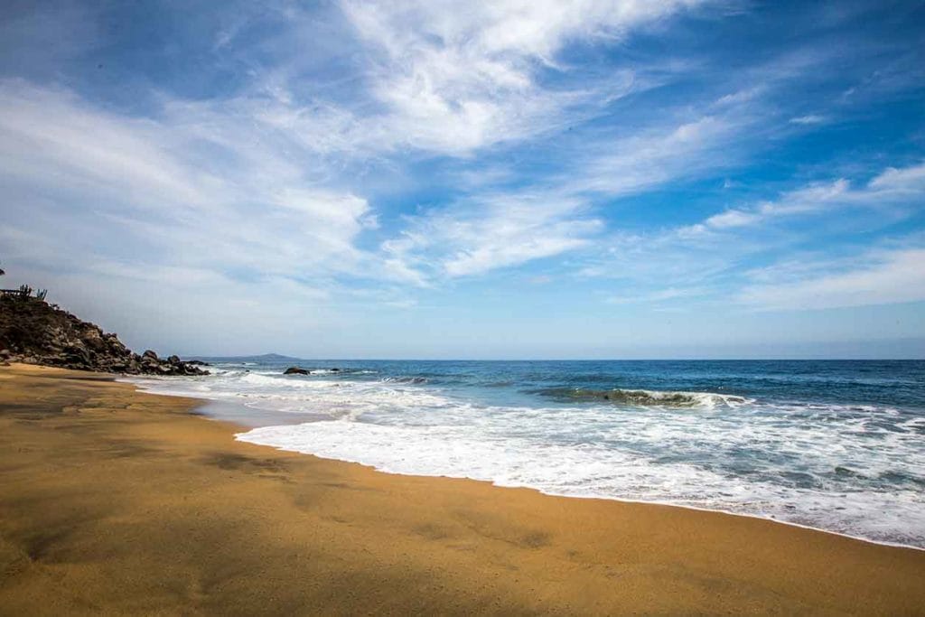 Peaceful beach scene in Sayulita with gentle waves rolling onto golden sand, cliffs in the distance, and a wide-open sky scattered with wispy clouds.