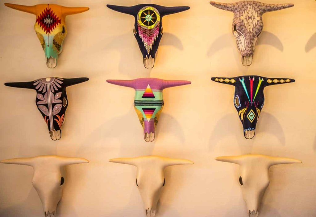 Wall display of nine painted bull skulls, each uniquely decorated with intricate patterns and bright colors, hanging on a cream-colored background.