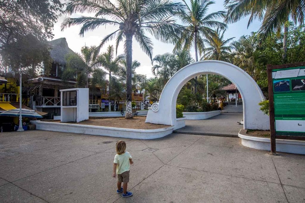 Little kid walking toward Sayulita’s town plaza, framed by palm trees and a white arched entrance, with shops and a quiet local vibe in the background.