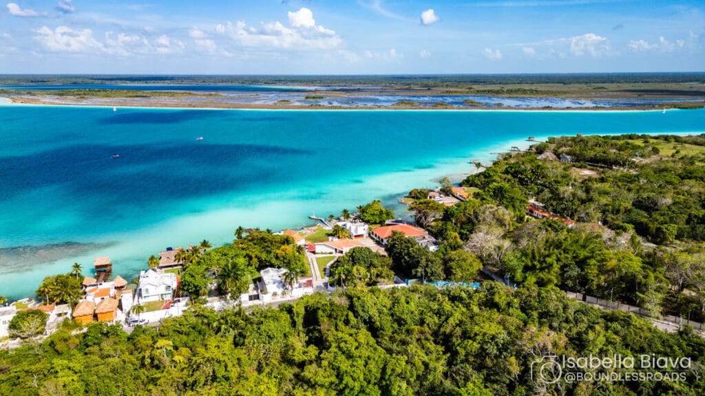 Aerial view of Bacalar Lagoon, Mexico. Turquoise water and lush greenery surround small buildings with red roofs, under a bright blue sky.