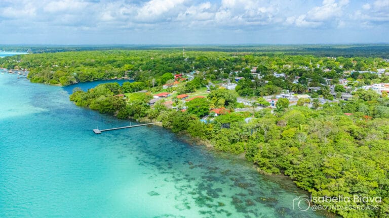 Aerial view of Bacalar, Mexico, showcasing lush greenery, turquoise lagoon, small dock, and scattered colorful houses under a blue sky with clouds.