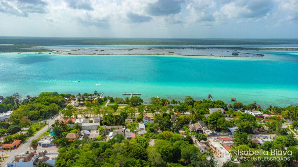 Aerial view of Bacalar Lagoon, Mexico, featuring turquoise water, lush greenery, small buildings, and boats, under a partly cloudy sky.