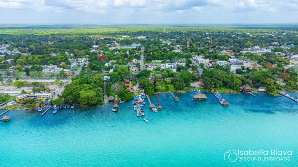 Aerial view of Bacalar, Mexico showcasing vibrant turquoise waters, lush greenery, docks, and distant buildings under a partially cloudy sky.