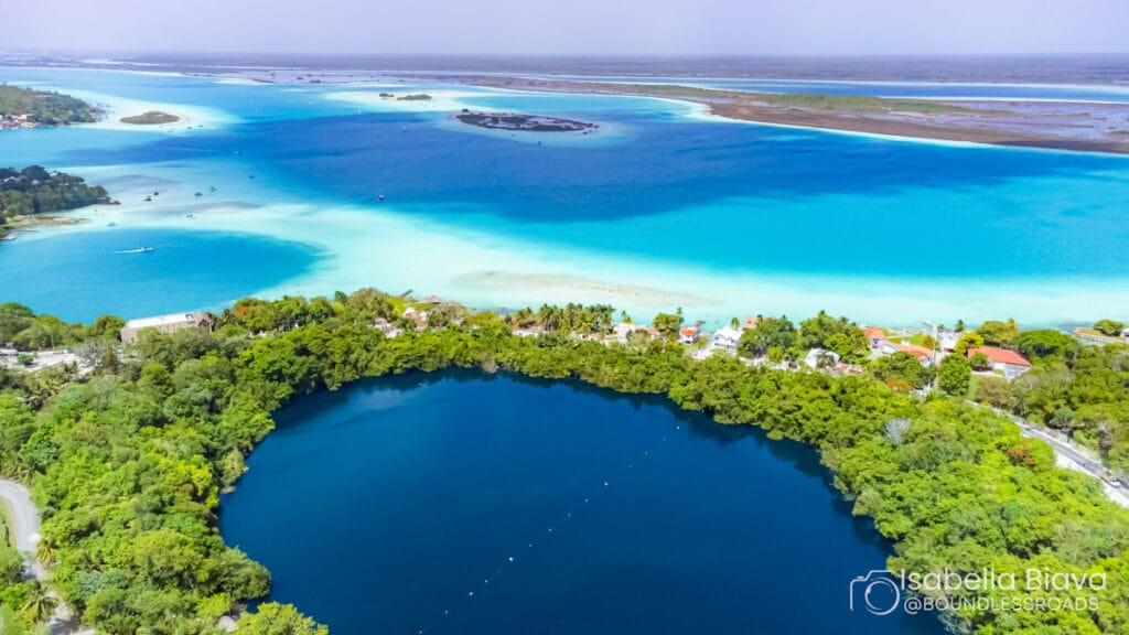 Aerial view of Mexico's Bacalar Lagoon featuring turquoise waters, a deep cenote, lush greenery, small buildings, and distant boats.