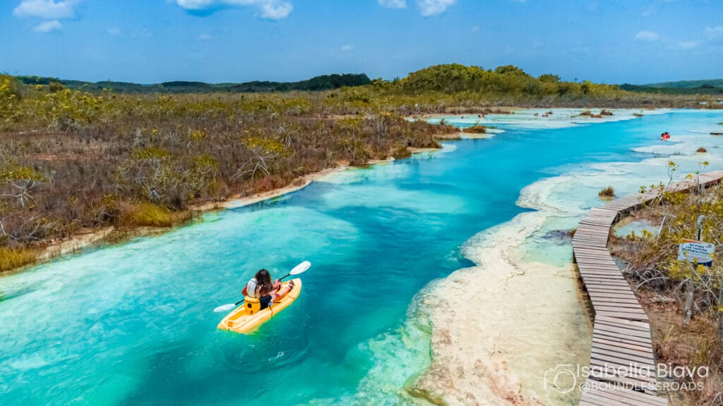 A person kayaking on a bright turquoise river surrounded by lush vegetation, with a wooden walkway meandering alongside the water.