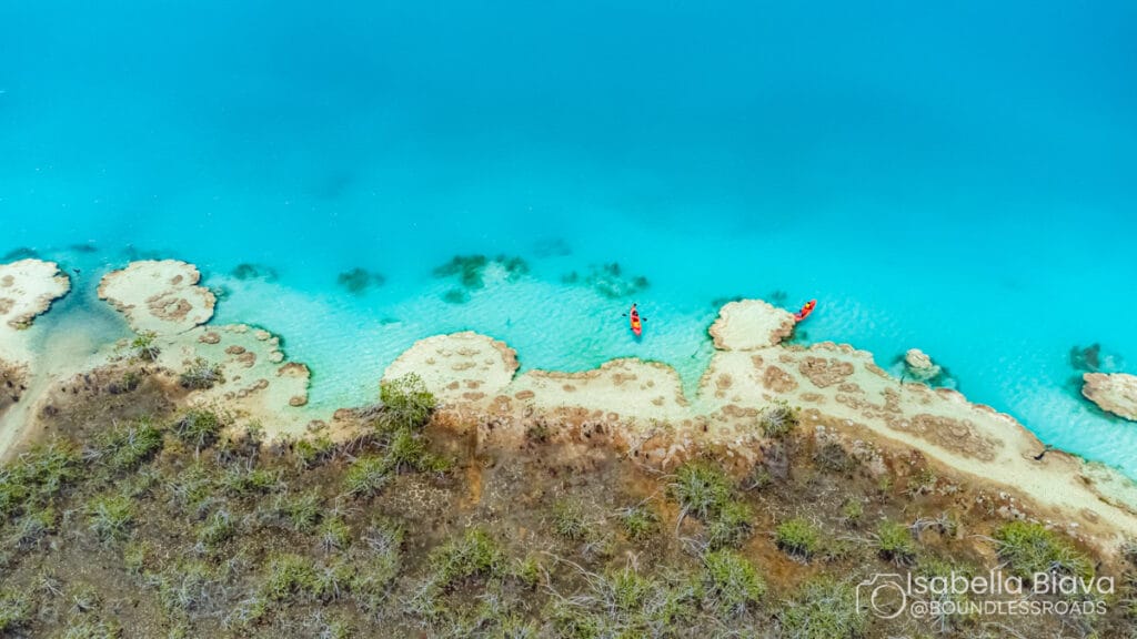 Aerial view of a person kayaking in a bright turquoise lake near a rugged shoreline with sparse vegetation and sandy formations.