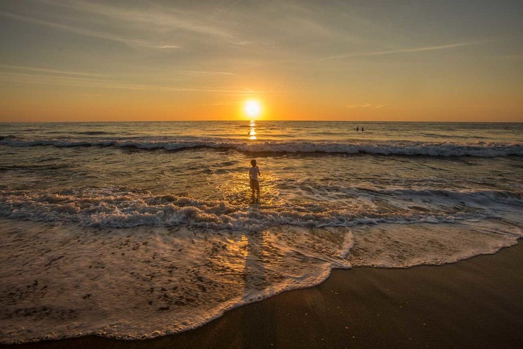 Golden sunset over the ocean with soft waves lapping the shore, and a child standing in the water casting a long shadow in the golden light.