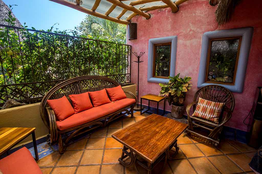 Cozy, rustic-style patio with terracotta tile floors, a bamboo roof, and cushioned rattan furniture set against a pink stucco wall and leafy backdrop.