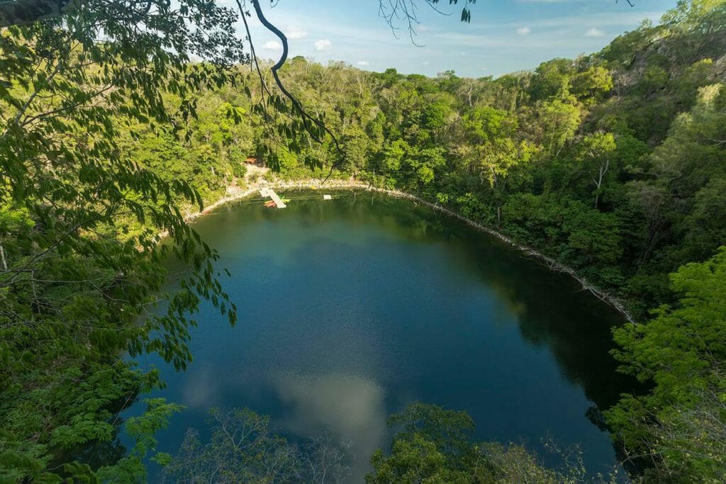 Aerial view of a serene, circular cenote surrounded by lush green forest, with a small platform at the edge.