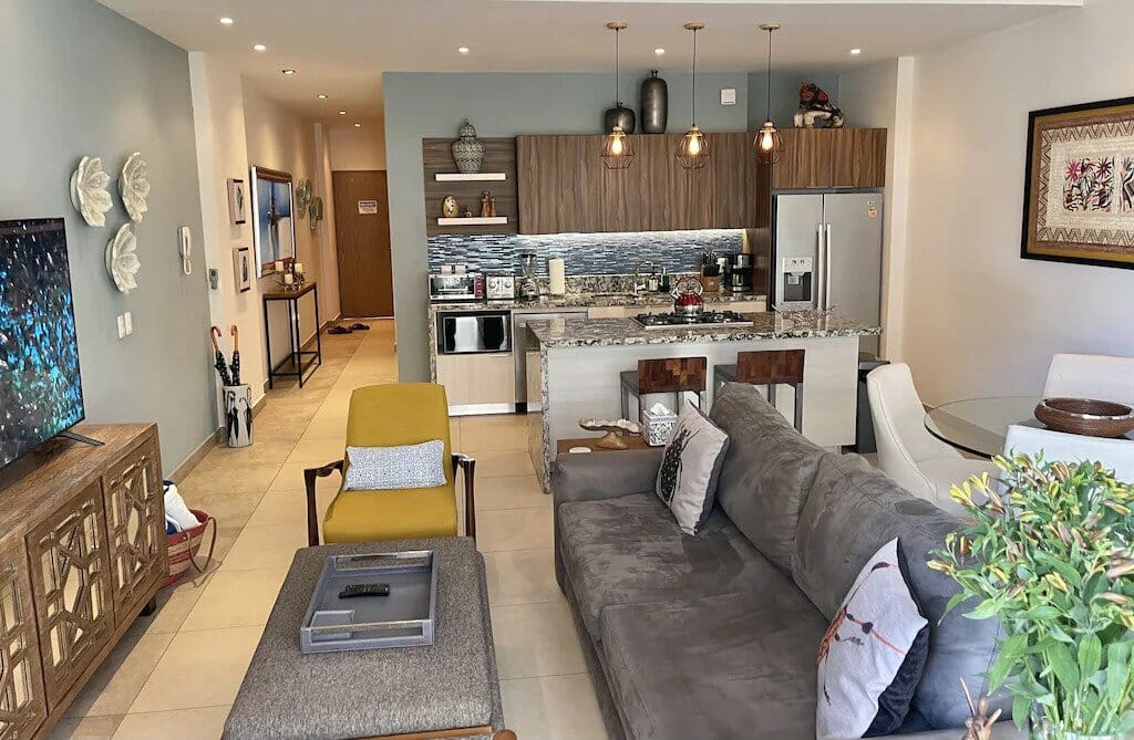 Modern living room and kitchen with wood accents, gray sofa, yellow chair, dining area, pendant lights, and decorative elements, creating a cozy, stylish atmosphere.