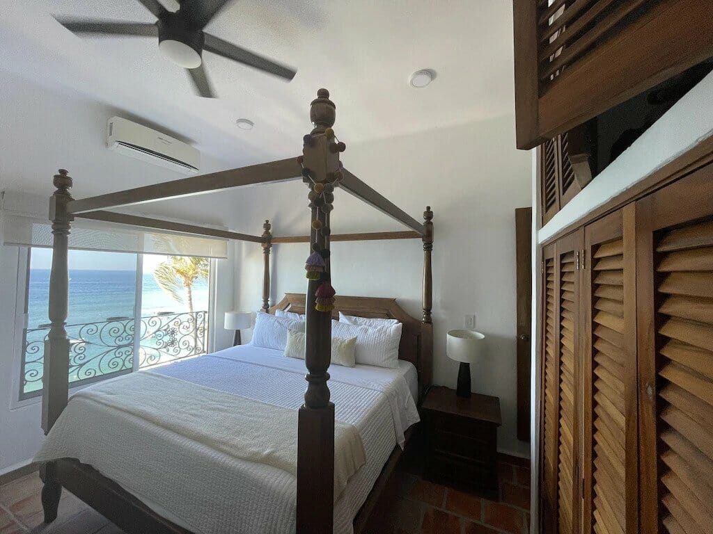 A serene bedroom with a canopy bed, ocean view, ceiling fan, wooden shutters, and cozy decor, overlooking the sea from a balcony.