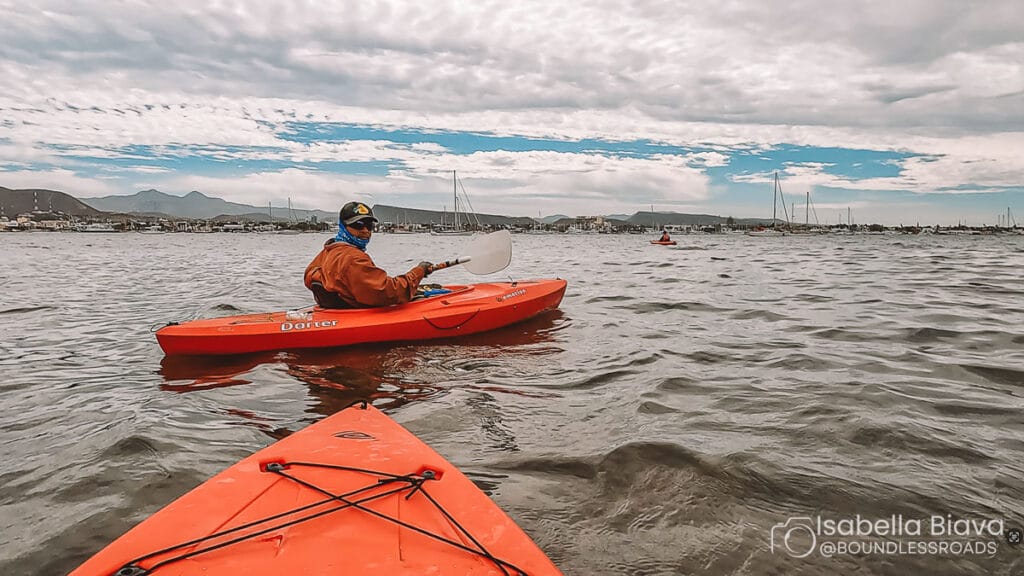 A person kayaking in a red kayak on a calm bay, surrounded by mountains and a marina filled with sailboats under a cloudy sky.