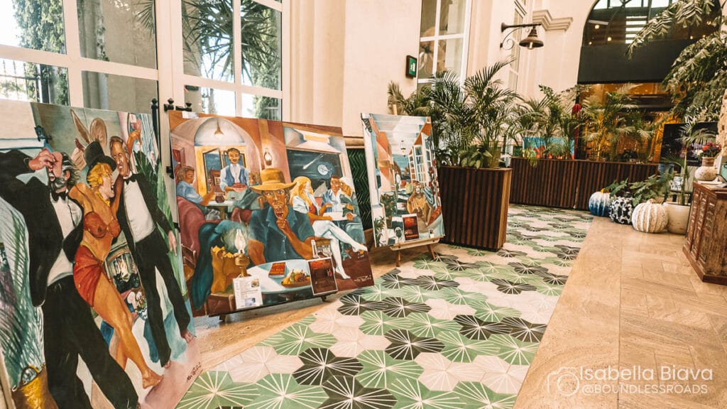Paintings displayed on patterned floor near large windows and plants in modern indoor setting. Vibrant artwork featuring lively scenes and figures.