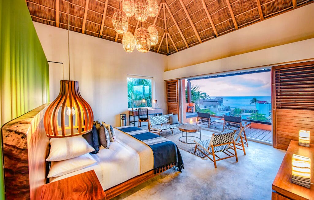 Bright and modern hotel room with a tropical flair, featuring a thatched ceiling, woven pendant lights, a king-sized bed, and a seating area with mid-century modern furniture. Large sliding doors open to a private balcony with ocean views and a coastal village below, glowing softly at sunset.