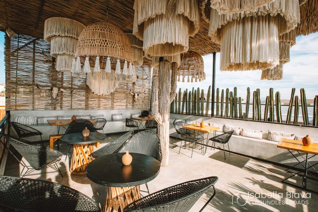 Chic outdoor café with wicker lamps, black tables, and woven chairs. Wooden accents and cacti fence create a rustic, serene atmosphere. No people visible.