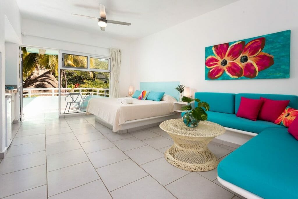 Bright and tropical suite with turquoise and white decor, featuring a bed, bold flower wall art, and a matching sofa with colorful pillows. Glass doors open to a private patio with palm views.