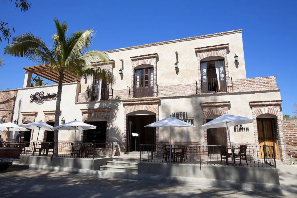 A two-story colonial-style building with brick arches, outdoor seating, and palm trees under a clear blue sky, labeled "Hotel Boutique."