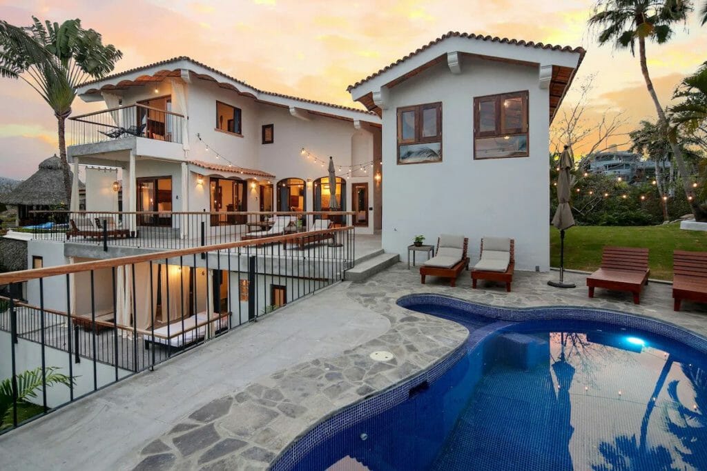 Chic white villa with a curved-edge pool, stone-tiled deck, and string lights glowing at sunset. Multiple balconies, cozy loungers, and palm trees complete the luxurious yet laid-back vibe.