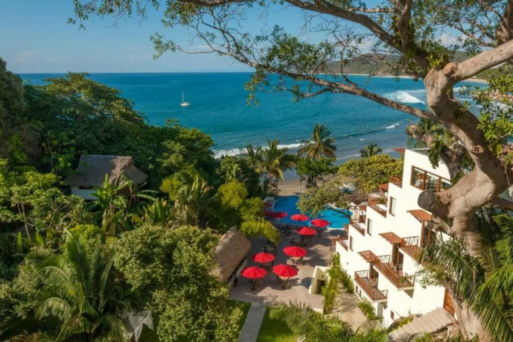 Oceanfront hotel surrounded by lush greenery and tropical trees, with red umbrellas dotting the pool area below. The beach and blue sea stretch out just steps away, creating a dreamy coastal escape.