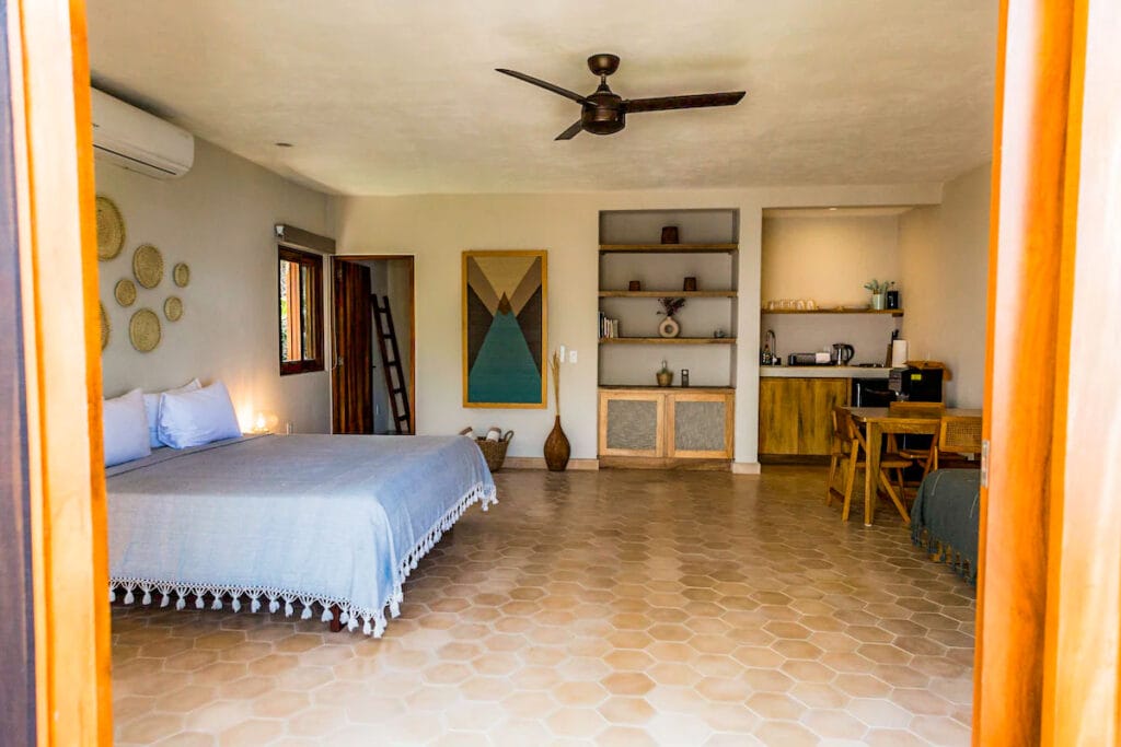 Bright, spacious bedroom with minimalist boho decor, featuring a king-sized bed, natural woven accents, open shelving, and a small kitchenette. A large window and neutral tones create an airy, serene atmosphere.