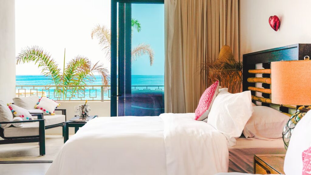 Bright and airy hotel room with a plush bed, bamboo headboard, and cozy seating area decorated with colorful pom-pom pillows. Sliding glass doors open to a private balcony with palm trees and a stunning ocean view just beyond the railing.
