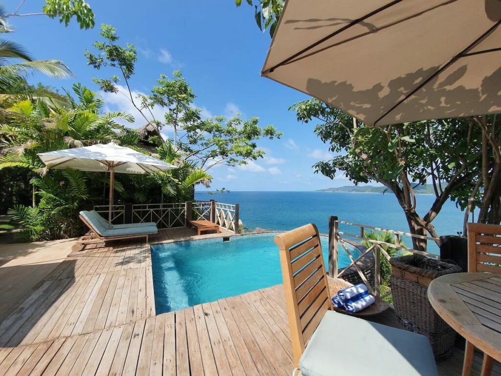 Ocean-view deck with a small infinity pool, wooden lounge chairs, and umbrellas nestled among lush trees. Overlooks a stunning blue bay, offering an ultra-private and peaceful setting.