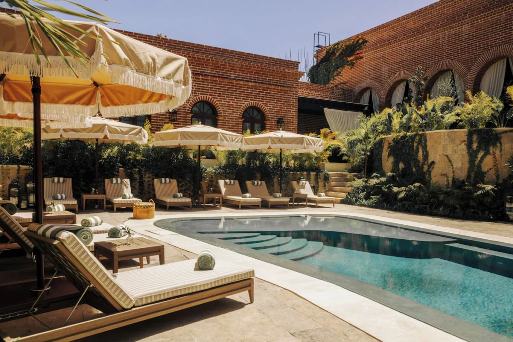 Charming courtyard with pool, loungers, and parasols, surrounded by lush greenery and brick architecture, creating a relaxing retreat atmosphere.