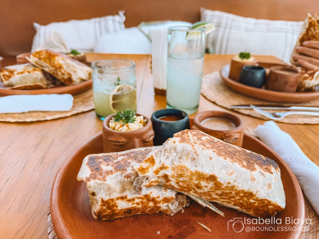 A cozy table setting with filled wraps, sauces, and drinks on wooden plates. Cushions in the background create a warm atmosphere.