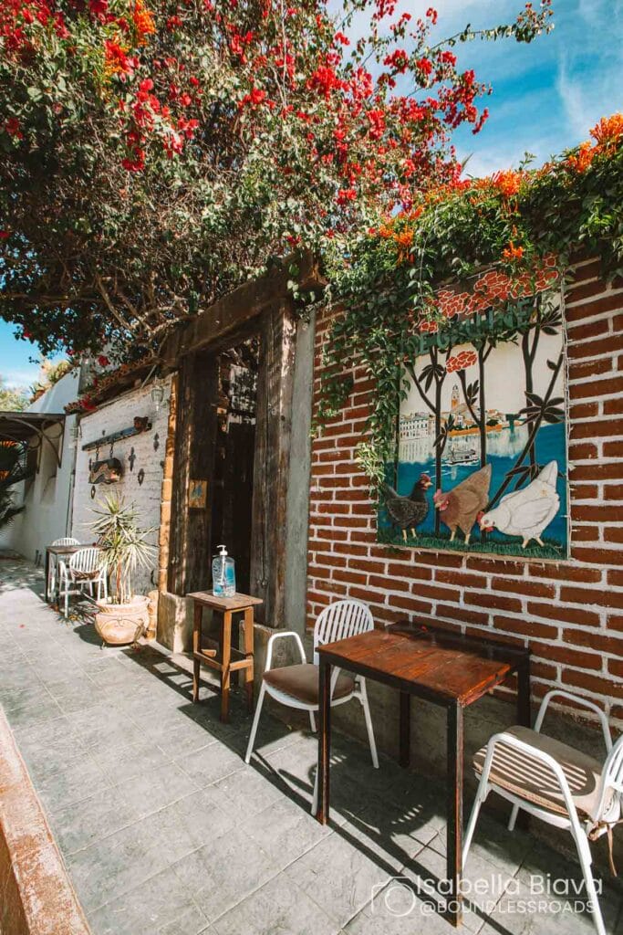 A charming outdoor cafe with colorful flowers, brick walls, and a whimsical mural featuring chickens. Tables and chairs line the shaded walkway.