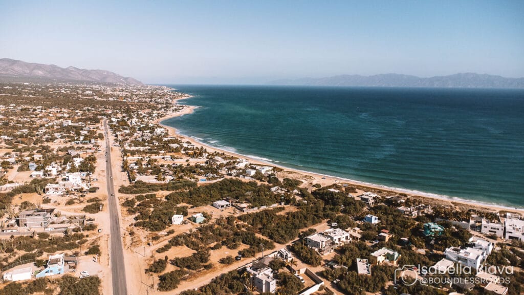 Coastal landscape features sandy beach, scattered buildings, and a winding road alongside the ocean, with distant mountains under a clear blue sky.