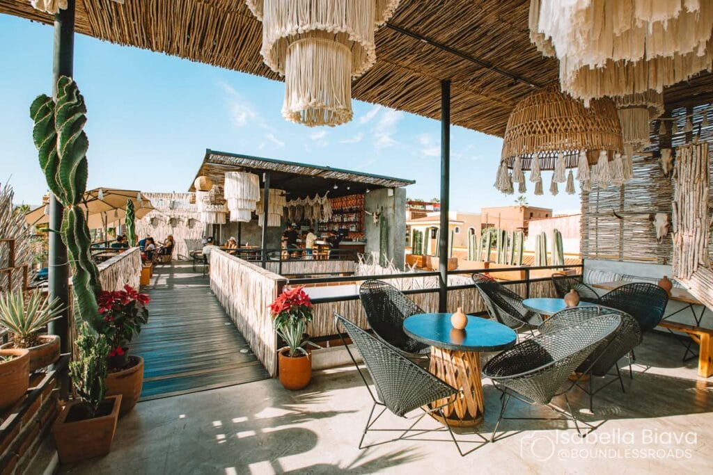 Rustic outdoor cafe with woven decor, tables, and chairs. People sitting, cacti, and red flowers create a warm, inviting atmosphere under clear blue sky.