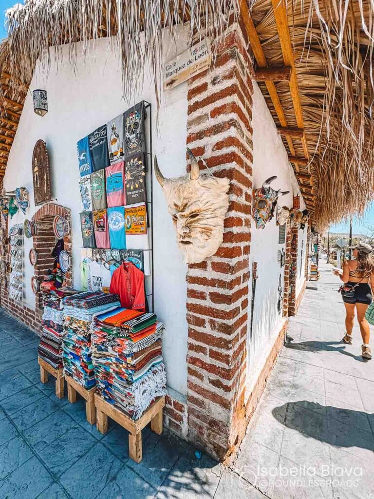 A street market corner displaying colorful textiles, T-shirts, and masks. A person walks by under a thatched roof on Calle Marquez de Leon.