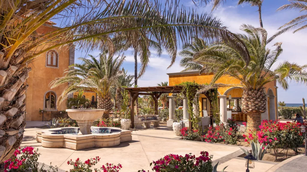 Tropical courtyard with palm trees, vibrant flowers, and a central fountain. Rustic building and pergola offer shaded seating, ocean visible in the background.
